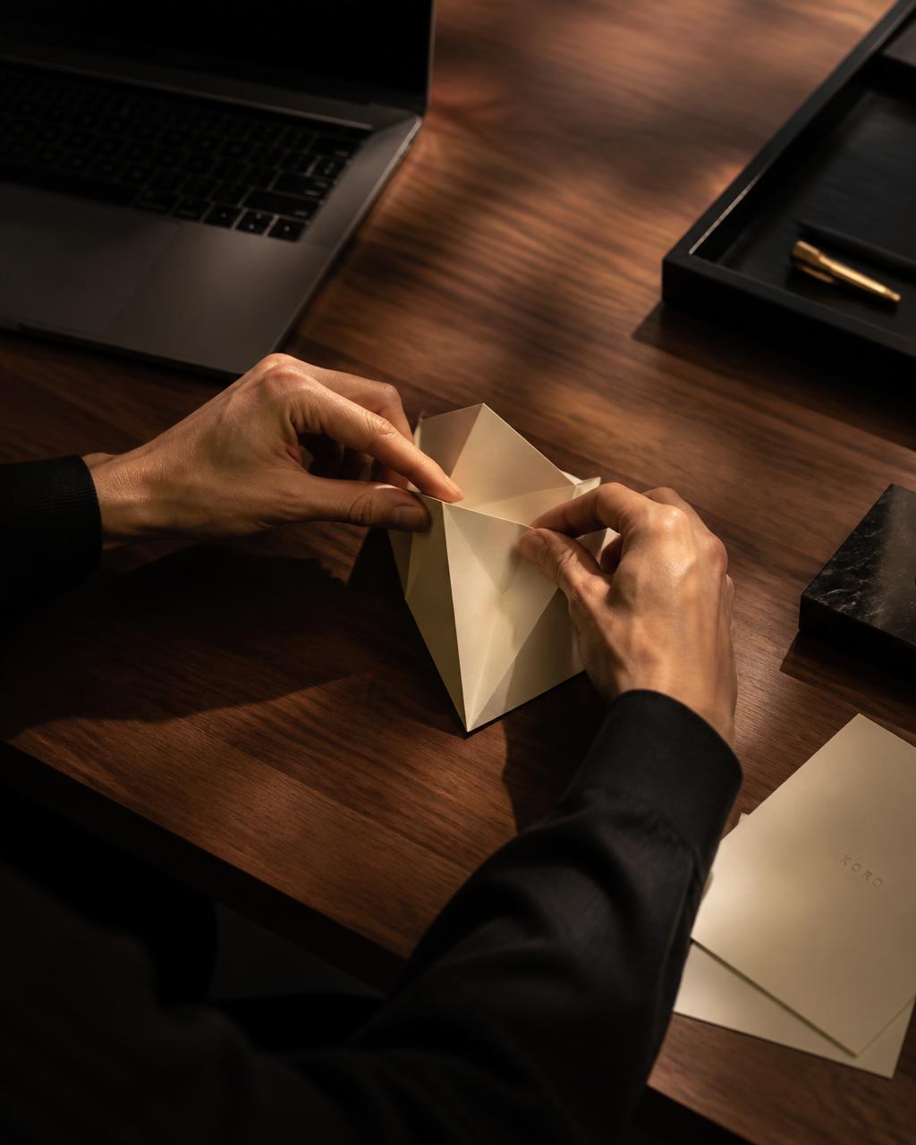 Hands folding a premium matte card into a geometric origami form on a walnut desk